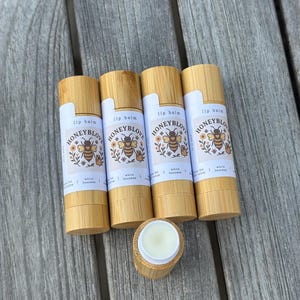 May include: Four lip balm tubes with bamboo-colored casings and white labels. The labels feature a bee illustration with the text "HONEYBLOVE" and "lip balm." One tube is open, revealing the balm. The background is a weathered wooden surface.