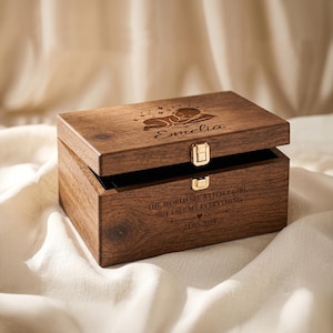 May include: A rectangular dark brown wooden keepsake box with a gold-tone metal latch. The lid features an engraved sleeping baby and the name Emelia. The front is engraved with the text: THE WORLD SEE A LITTLE GIRL, BUT I SEE MY EVERYTHING 21.05.2024.