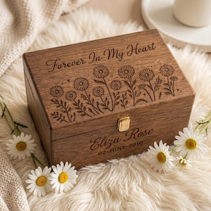 May include: A wooden box with the inscription "Forever In My Heart" and floral engravings. The box features a gold-colored clasp and the name "Eliza Rose" with the date "02-JUNE-2010". The box is surrounded by white daisies.
