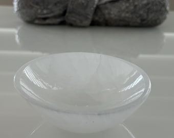 Puede incluir: Un cuenco pequeño, blanco y translúcido, de forma redondeada. El cuenco está hecho de un material liso y pulido y refleja la luz. En el fondo se ve una estatua de una figura sentada.
