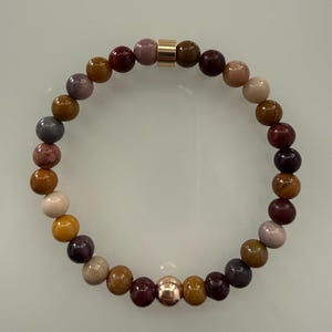 May include: A beaded bracelet featuring a circular arrangement of smooth, multi-coloured stone beads in shades of brown, tan, mauve, and deep burgundy. The design includes a single polished gold-tone cylindrical bead and a round rose gold-tone bead.