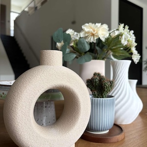 以下が含まれることがあります： 木製の表面に、装飾用の花瓶と小さな鉢植えのサボテンが並んでいます。花瓶には、ベージュのドーナツ型花瓶、花が入ったピンクの花瓶、白のテクスチャード加工された花瓶があります。サボテンは青いストライプの鉢に入っています。