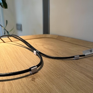 Pode incluir: Cabos pretos com clipes brancos em uma superfície de madeira. Os cabos são curvos e parecem fazer parte de um dispositivo eletrônico. O fundo é uma parede de cor clara e uma planta.