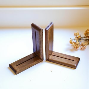 May include: Two wooden bookends in a warm brown hue. Each bookend has an L-shape with a vertical support and a horizontal base. A groove runs along the base. The bookends are set against a white background.