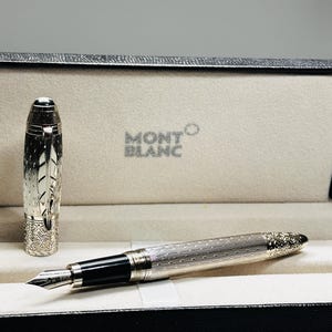 May include: A silver Montblanc fountain pen with intricate engraved detailing, presented in an open black box with a cream-colored interior. The pen features a black nib and a cap with a decorative design. The Montblanc logo is visible.