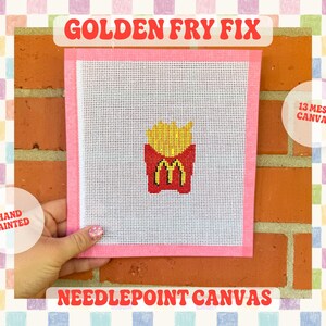 Puede incluir: Un lienzo cuadrado de punto de aguja con un gráfico pintado a mano de papas fritas amarillas en un recipiente rojo con un logo M amarillo. El lienzo tiene un borde rosa y está marcado como 13 mesh canvas. El texto GOLDEN FRY FIX y NEEDLEPOINT CANVAS aparece.