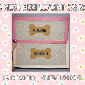 May include: Hand-painted needlepoint canvas with a pink border. The canvas features two dog bone designs with the names "Archie" and "Duncan". The text "18 MESH NEEDLEPOINT CANVAS" and "HAND PAINTED | CUSTOM DOG BONE" are also visible.