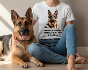 May include: A white t-shirt with a graphic of a German Shepherd dog and the text "SECURITY SYSTEM: ARMED, LOUD, AND SHEDDING." The dog is sitting next to a person wearing blue jeans.