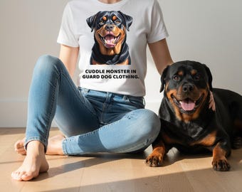 May include: White t-shirt with a Rottweiler graphic and the text "Cuddle Monster in Guard Dog Clothing." A Rottweiler dog is lying on the floor next to the person wearing the shirt. The dog has black and brown fur.