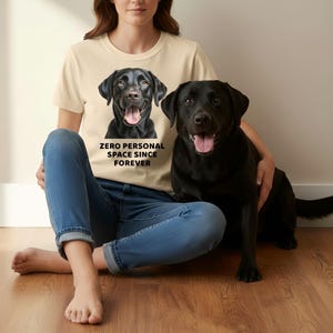 May include: A cream-colored t-shirt featuring a graphic of a black Labrador Retriever with the text "ZERO PERSONAL SPACE SINCE FOREVER." A black Labrador Retriever sits next to the person wearing the shirt.