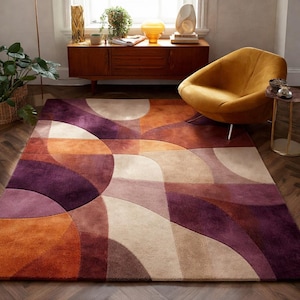 Könnte beinhalten: Ein großer Teppich mit geometrischem Design in Lila-, Orange- und Beigetönen. Der Teppich weist geschwungene und rechteckige Formen auf, die ein modernes und künstlerisches Muster erzeugen. Der Teppich befindet sich in einem Wohnzimmer mit einem senffarbenen Stuhl.