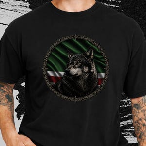 May include: Black t-shirt featuring a detailed illustration of a wolf inside a circular frame. The frame surrounds a green and red flag design. The wolf is depicted with dark fur and a focused gaze, creating a striking contrast against the black fabric.