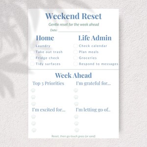 May include: A white paper with the text "Weekend Reset" in a soft blue font. The paper is divided into sections for home, life admin, and week ahead tasks. Includes prompts for gratitude and priorities.