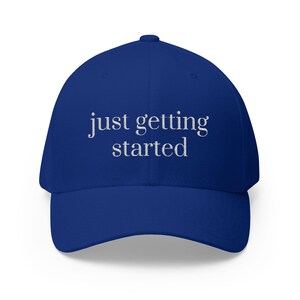 May include: A royal blue baseball cap with the phrase "just getting started" embroidered in white. The cap has a curved brim and a structured fit, suitable for casual wear.