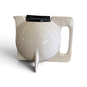 May include: A cream-colored ceramic teapot with a spherical body, a single pointed base support, and a rectangular handle. The teapot features a flat, black lid positioned on top, creating a distinct contrast against the light-colored ceramic surface.