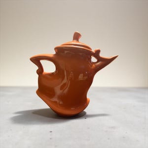 May include: A glossy, orange ceramic teapot with a unique, abstract design. The teapot features a curved handle, a pointed spout, and a lid with a small knob. The piece is set against a neutral background, highlighting its sculptural form.