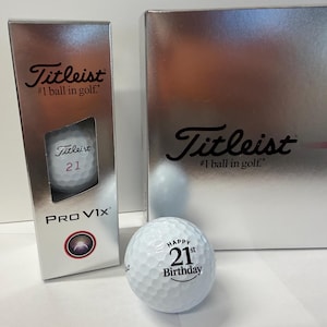 May include: A white Titleist Pro V1x golf ball with the text Happy 21st Birthday printed on it, positioned in front of a silver vertical box and a larger silver box, both featuring the Titleist #1 ball in golf logo and branding.
