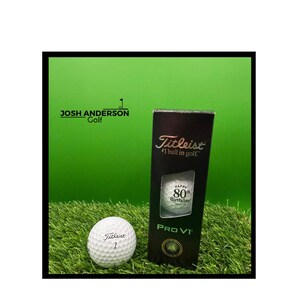 May include: A white Titleist Pro V1 golf ball with the number 1 printed on it, positioned next to a black vertical box. The box features the Titleist logo, the text #1 ball in golf, PRO V1, and a golf ball printed with Happy 80th Birthday.