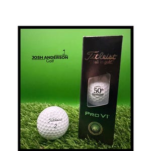 May include: A white Titleist Pro V1 golf ball with the number 2 printed on it, alongside a black vertical box containing another golf ball with the text Happy 50th Birthday printed on it. The box also features the Titleist logo and text.