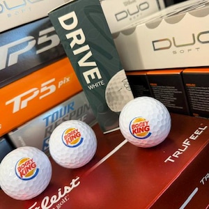 May include: Three white golf balls featuring a circular Bogey King logo printed in orange, blue, and red. The balls are arranged in front of a collection of stacked golf ball boxes from brands such as TaylorMade, Wilson, and Titleist, plus a Vice Drive box.