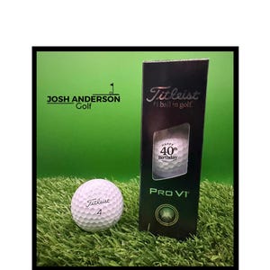 May include: A white Titleist Pro V1 golf ball marked with the number 4 and a matching black box. The box features the Titleist logo, the text #1 ball in golf, and a window displaying a golf ball printed with the text Happy 40th Birthday.