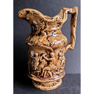 May include: A brown ceramic pitcher with a handle and a spout. The pitcher is decorated with relief carvings of grapes, leaves, and figures in classical attire. The pitcher has a pedestal base and is likely vintage.