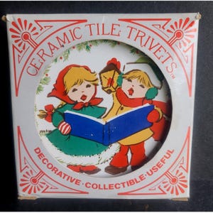 May include: A ceramic tile trivet in a box. The trivet features a colorful illustration of two children singing from a blue book, one holding a lantern. The box reads "CERAMIC TILE TRIVETS" and "DECORATIVE COLLECTIBLE USEFUL".