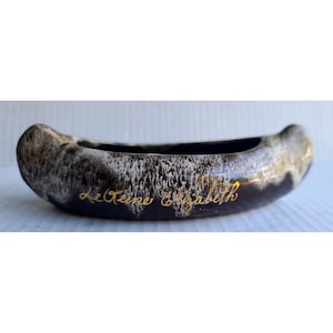 May include: A canoe-shaped ceramic planter featuring a dark brown and white drip glaze finish. The exterior is decorated with the gold cursive text Le Reine Elizabeth. The vessel has a curved, elongated form with raised ends and a smooth, glossy surface.