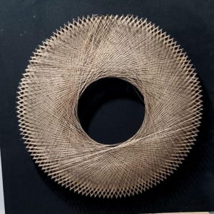 May include: A circular string art piece with a central hole. The artwork features a woven pattern of beige string, creating a textured, radial design. The piece is set against a black background, highlighting the intricate details of the string arrangement.