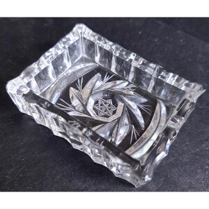 May include: A clear, rectangular glass ashtray with a decorative, starburst pattern in the center. The ashtray has a faceted edge and is made of thick, clear glass. The design features a central star and radiating lines.