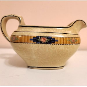 May include: Cream-colored ceramic creamer with a handle and spout. The creamer features a decorative band with orange stripes, blue geometric shapes, and pink rose accents. Black trim outlines the top and base.