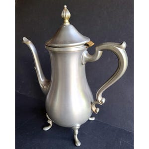 May include: A silver-colored metal coffee pot with a curved spout, handle, and lid. The lid is topped with a gold-colored finial. The pot has three small feet and a decorative handle. The background is black.
