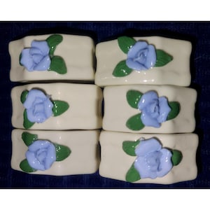 May include: Six off-white ceramic napkin rings, each adorned with a light blue rose and green leaves. The floral design is centered on the top of each rectangular ring, creating a cohesive set for table settings.