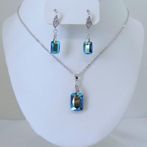 May include: A silver-toned jewelry set featuring a necklace and matching earrings. The set includes a pendant and two earrings, each with a rectangular, iridescent blue-green crystal. The earrings dangle from decorative silver hooks.