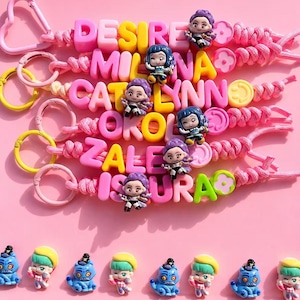 May include: Keyrings with names spelled out in pink and yellow letter beads. Each keyring has a small doll charm. Additional doll charms are lined up at the bottom of the image. The background is pink.