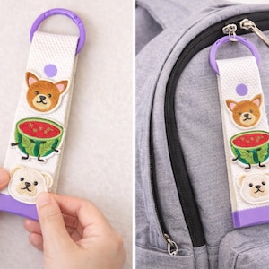 May include: White fabric keychain with a purple loop and base, adorned with embroidered patches of a Shiba Inu dog, a watermelon, and a bear. The keychain is attached to a gray backpack.