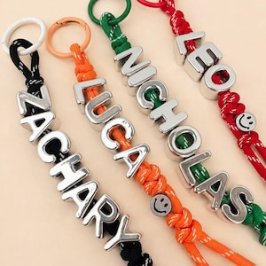 Op de afbeelding: Vier gepersonaliseerde sleutelhangers met gevlochten nylon koorden in zwart, oranje, groen en rood. Elk koord is bevestigd aan een bijpassende kunststof ring en voorzien van zilverkleurige blokletters die ZACHARY, LUCA, NICHOLAS en LEO spellen, plus smiley-kralen.