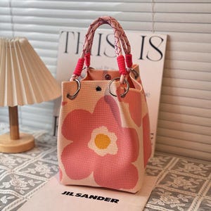 May include: A structured tote bag featuring a large pink and yellow floral pattern on a light peach background. The bag has two braided rope handles with bright pink accents, silver-tone metal rings, and a central snap closure at the top opening.
