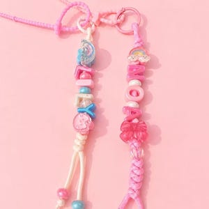 May include: Two beaded phone charms featuring pink and blue colour schemes. One charm spells LILY with a rainbow and cloud bead, while the other spells NOA with a rainbow, cloud, and pink bow bead. Both charms include various plastic beads.