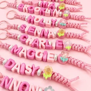 May include: A collection of pink braided cord keychains featuring custom names spelled out in pink and white plastic letter beads, accented with small white spherical beads and translucent flower-shaped charms in various pastel colours.