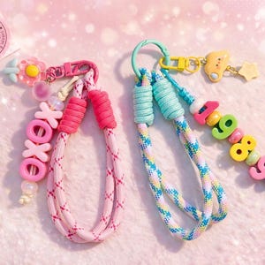 May include: Two wristlet keychains made of braided cord. The left keychain is pink with a pink XOXO bead charm and a flower charm. The right keychain is blue and white with a yellow cloud charm, a star charm, and colorful beads spelling 1989.