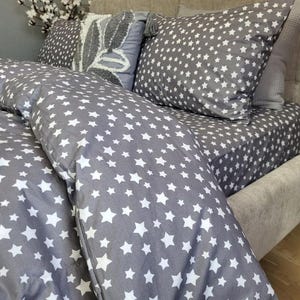 May include: A grey bed set featuring a white star design. The set includes a duvet cover, pillowcases, and a fitted sheet. Decorative pillows in shades of grey complement the bedding. The bed is upholstered in a neutral fabric.