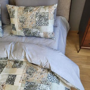 Könnte beinhalten: Ein Bett mit hellblauer Bettdecke und zwei dekorativen Kissen. Die Kissen und der untere Teil der Bettdecke weisen ein Patchwork-Design in Beige-, Grau- und Weißtönen auf. Im Hintergrund ist eine Holzkommode zu sehen.