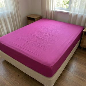 May include: A solid magenta fitted sheet covering a mattress. The fabric is smooth and stretched taut across the surface, displaying a vibrant pinkish-purple hue that covers the entire top area and sides of the bed frame in a clean, uniform manner.