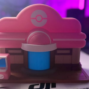 May include: A pink and brown plastic model of a Pokemon Center building featuring a white Poke Ball emblem on the roof, a central blue doorway, and a small pink arcade-style machine positioned to the left on a brown rectangular base.