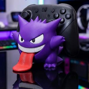 May include: A purple Gengar character figure serving as a controller stand, featuring a wide grin with white teeth and a long red tongue extending downward, holding a black wireless gaming controller behind its back on a reflective surface.