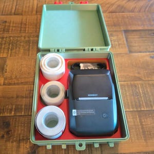 May include: A portable black Niimbot label printer with a power button and a paper feed slot, housed in an open olive green plastic carrying case. The case includes three white rolls of adhesive label tape and a black charging cable in red slots.