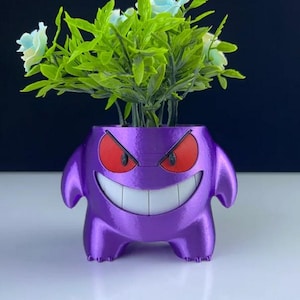 May include: A purple planter shaped like a cartoon character with red eyes and a wide grin, holding green foliage and light blue flowers. The planter has small arms and feet, and is a unique decorative item.
