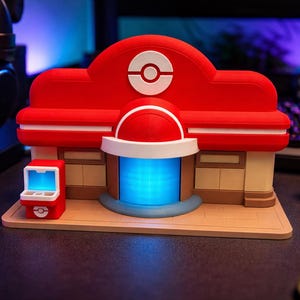 May include: A red and white building model with a blue glowing entrance and a small red arcade machine. A red and white ball sits to the right. The model is on a beige base.