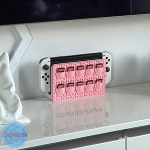 May include: A pink 3D-printed game cartridge holder with a basket weave design, holding multiple game cartridges. The holder is designed to fit a Nintendo Switch console, which is also visible in the image.